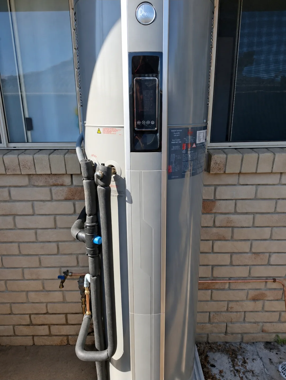 A Rheem heat pump hot water system installed by RCM against a brick wall, controller visible