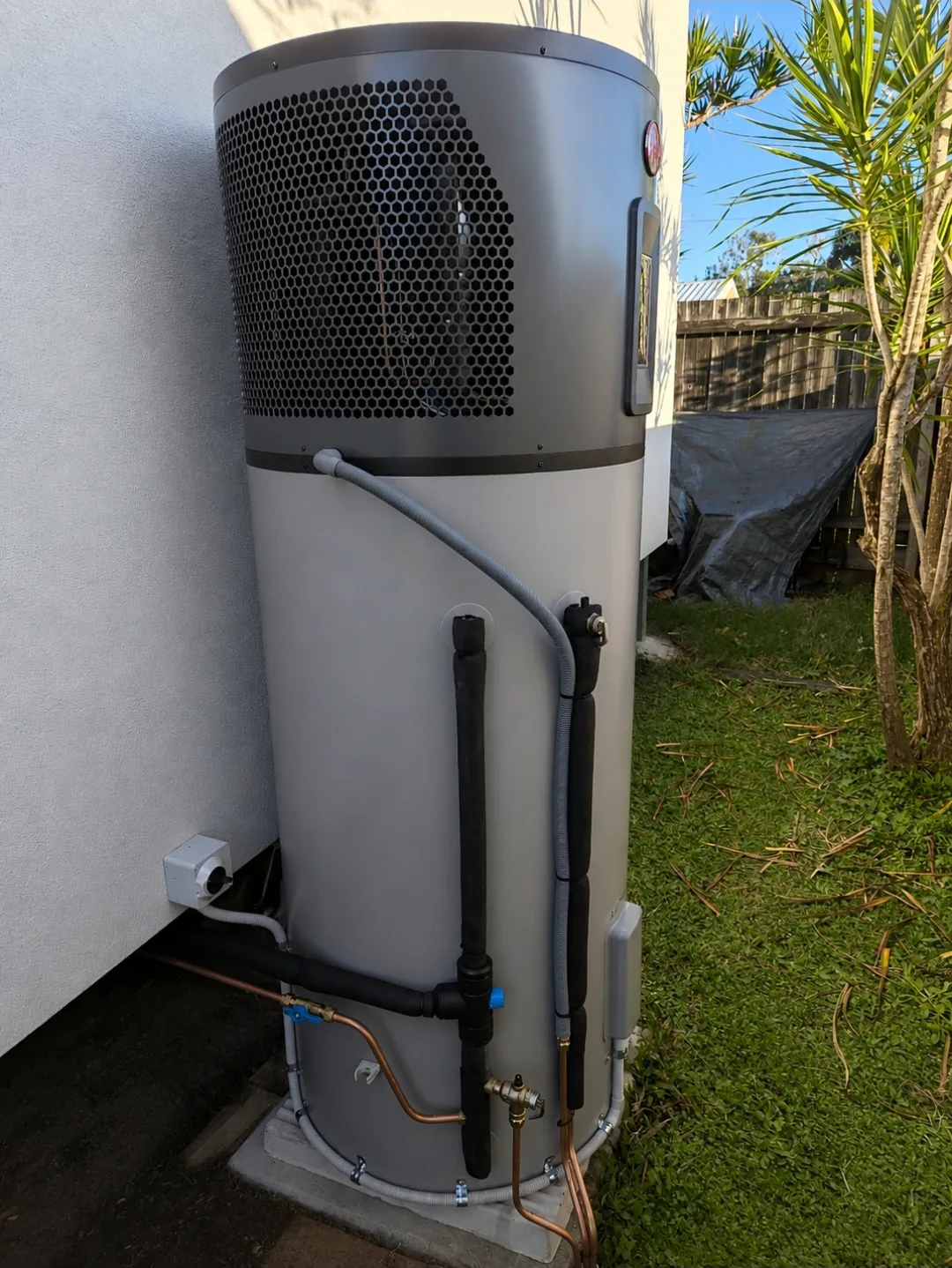 A Rheem heat pump hot water system installed by RCM against a house wall, showing clearance and condensate drainage